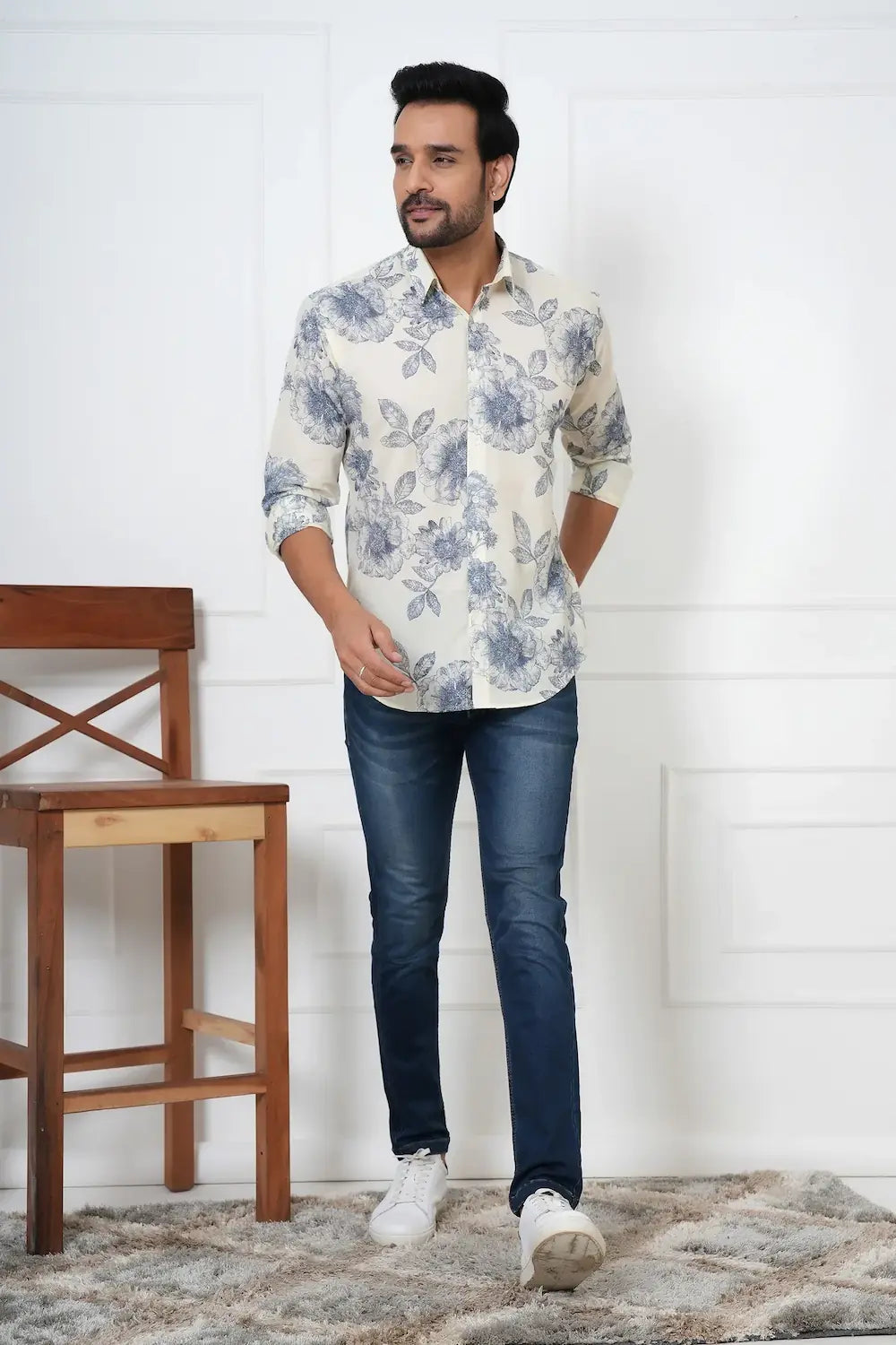 Man wearing a floral shirt and jeans standing in a room with wooden chair and white wall.